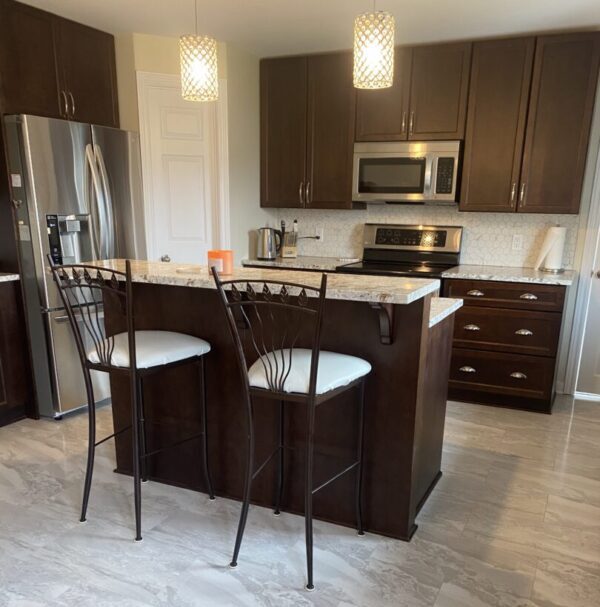 New build kitchen with a centre island and stainless steel appliances. Dark cabinets and light marble countertops. 2 barstools sit against the back of the island.