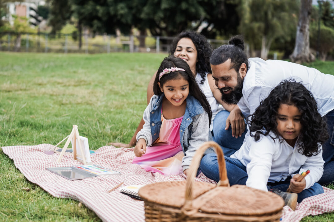 A family of four smiling sitting on a picnic blanket with a small easel and paints. A large picnic basket is in the foreground.